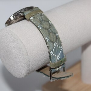 May include: A watch with a patterned strap featuring small white flowers on a blue-green background. The watch band is secured with a silver buckle. The watch is displayed on a white cylindrical stand.