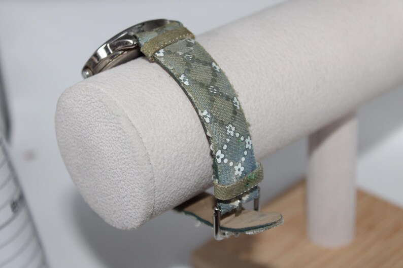 May include: A watch with a patterned strap featuring small white flowers on a blue-green background. The watch band is secured with a silver buckle. The watch is displayed on a white cylindrical stand.