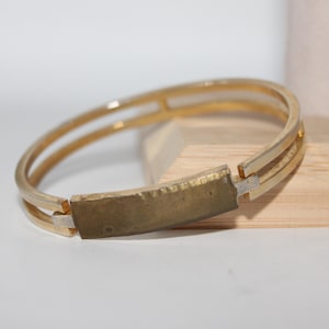May include: Gold-toned bangle bracelet with a rectangular center piece. The bracelet has a double-band design with a textured, brass-colored center. The bracelet is displayed on a light-colored surface.