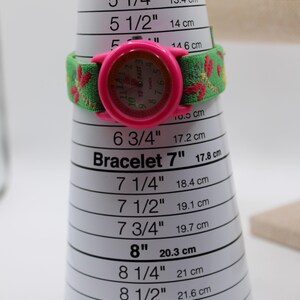 May include: A pink Timex watch with a green and red patterned strap sits on a bracelet sizing cone. The watch face is pink with black numerals. The cone measures from 12.7 cm to 22.2 cm, with markings in inches and cm.