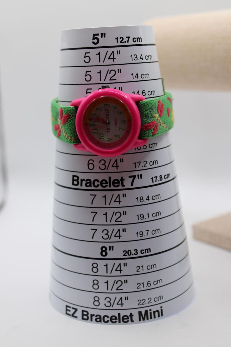 May include: A pink Timex watch with a green and red patterned strap sits on a bracelet sizing cone. The watch face is pink with black numerals. The cone measures from 12.7 cm to 22.2 cm, with markings in inches and cm.