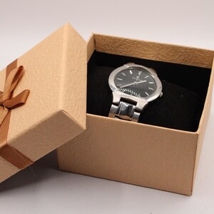 May include: A silver wristwatch with a black face rests inside an open, tan gift box. The box has a brown ribbon tied in a bow. The watch has a silver band and a black watch face with silver hands and markers. The box is lined with black fabric.