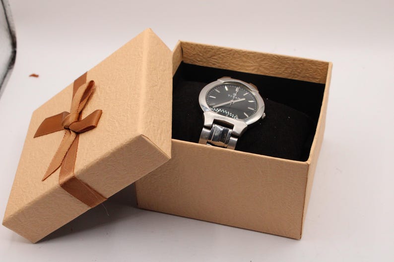May include: A silver wristwatch with a black face rests inside an open, tan gift box. The box has a brown ribbon tied in a bow. The watch has a silver band and a black watch face with silver hands and markers. The box is lined with black fabric.