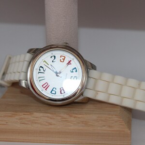 May include: A silver wristwatch with a white face and a white silicone band. The watch face has colorful numbers and hands. The watch is displayed on a wooden stand.
