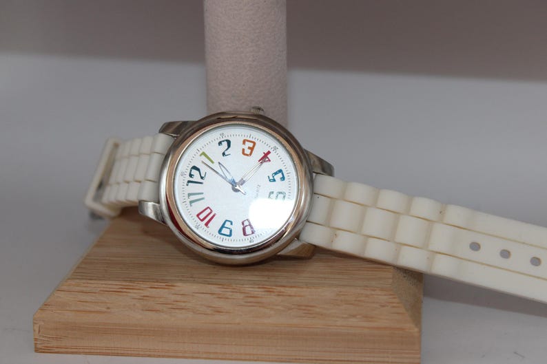May include: A silver wristwatch with a white face and a white silicone band. The watch face has colorful numbers and hands. The watch is displayed on a wooden stand.