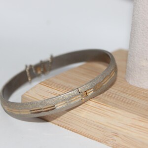 May include: A silver-toned hinged bangle bracelet with gold-colored accents. The bracelet is resting on a wooden surface next to a white cylindrical object. The bracelet has a textured finish and a rectangular gold design.