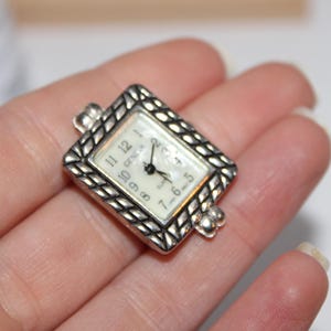 May include: A silver-toned Geneva watch with a rectangular face and a braided bezel. The watch face is white with black numerals and hands. The watch is held in a hand.
