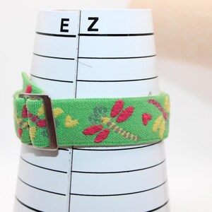 May include: A green elastic belt with a metal buckle, wrapped around a white cylindrical object. The belt is decorated with embroidered red dragonflies and yellow hearts. The object has black lines and the letters "E" and "Z" printed on it.