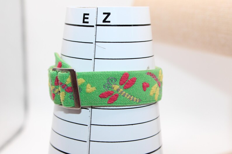 May include: A green elastic belt with a metal buckle, wrapped around a white cylindrical object. The belt is decorated with embroidered red dragonflies and yellow hearts. The object has black lines and the letters "E" and "Z" printed on it.
