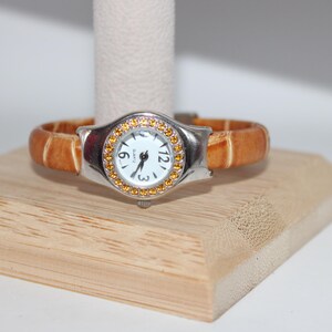 May include: A silver-toned wristwatch with a white face and gold-colored accents. The watch has a light brown leather band. The watch is displayed on a wooden stand.