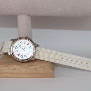 May include: A white wristwatch with a silver-colored case and a white band. The watch face is white with colorful numbers and hands. The watch is displayed on a wooden stand.