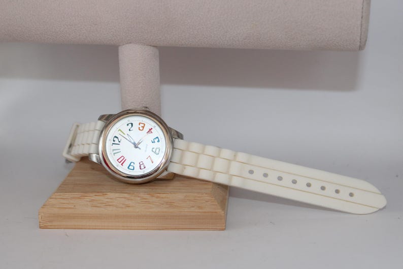 May include: A white wristwatch with a silver-colored case and a white band. The watch face is white with colorful numbers and hands. The watch is displayed on a wooden stand.