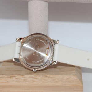 May include: A silver-toned watch with a white silicone band. The watch face is not visible. The back of the watch is visible, with text and a silver metal casing. The watch is resting on a wooden block.
