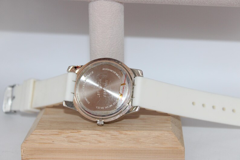 May include: A silver-toned watch with a white silicone band. The watch face is not visible. The back of the watch is visible, with text and a silver metal casing. The watch is resting on a wooden block.