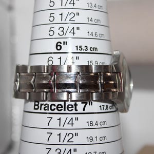May include: A silver-toned metal watch bracelet is measured on a white bracelet sizing cone. The cone indicates a bracelet size of 7 inches (17.8 cm). The watch face is visible on the right side.