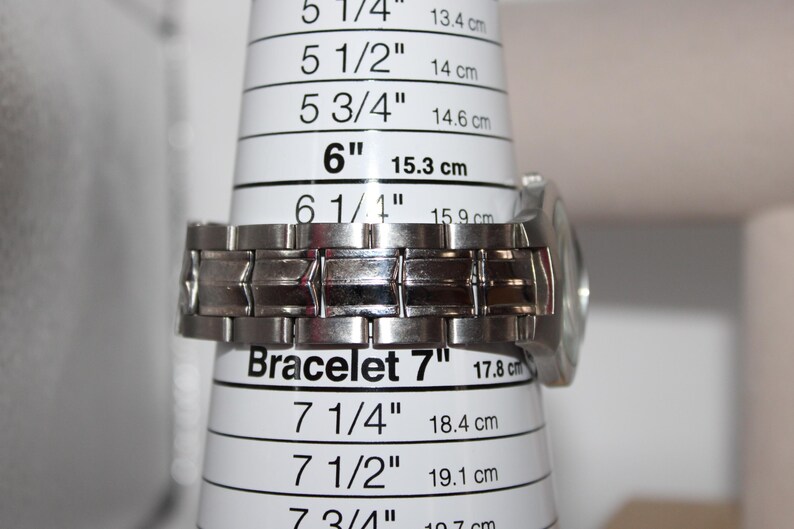 May include: A silver-toned metal watch bracelet is measured on a white bracelet sizing cone. The cone indicates a bracelet size of 7 inches (17.8 cm). The watch face is visible on the right side.