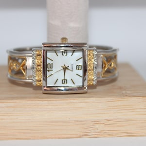 May include: A silver and gold-toned quartz watch with a rectangular face and a white dial. The watch features gold-colored accents and is displayed on a wooden stand. The watch has the numbers 12, 6, 9, and 3 on the face.