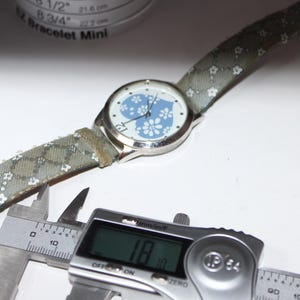 May include: A silver wristwatch with a light blue floral heart design on the face and a patterned strap. The watch is being measured with a digital caliper, showing a measurement of 1.8 inches.