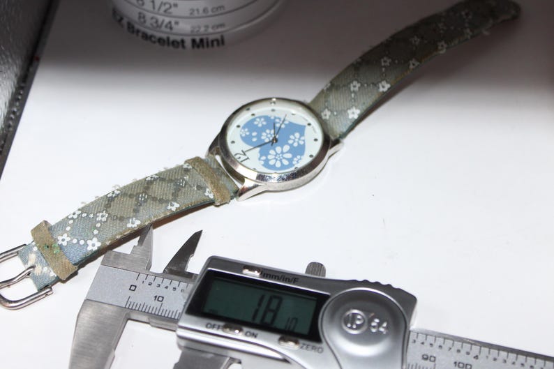 May include: A silver wristwatch with a light blue floral heart design on the face and a patterned strap. The watch is being measured with a digital caliper, showing a measurement of 1.8 inches.