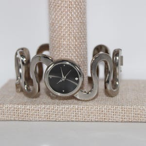 May include: A silver-toned wristwatch with a black face and a single diamond accent. The watch features a unique, wavy band design. The watch is displayed on a neutral-toned, textured surface.