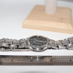 May include: A silver-toned wristwatch with a dark face and a stainless steel band. The watch is displayed on a white surface with a wooden block and a ruler showing inches. The watch band is approximately 4 inches long.