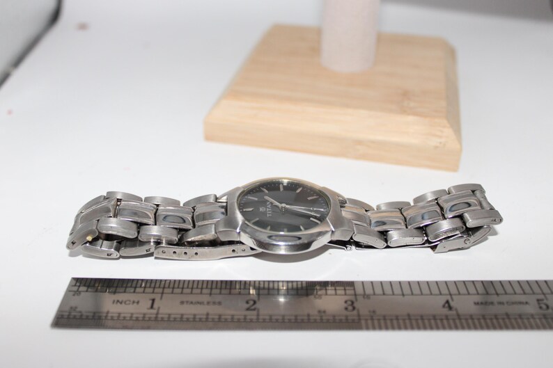 May include: A silver-toned wristwatch with a dark face and a stainless steel band. The watch is displayed on a white surface with a wooden block and a ruler showing inches. The watch band is approximately 4 inches long.
