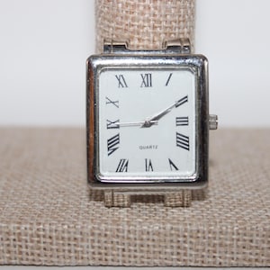 May include: Silver rectangular quartz watch with a white face and black Roman numerals. The watch has a silver band and is displayed on a beige surface. The watch hands are black.