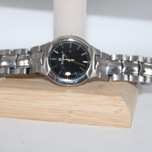May include: A silver-toned Titan wristwatch with a black watch face. The watch has a metallic band and is displayed on a wooden stand. The watch face has silver hour markers and hands.