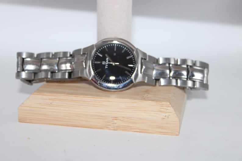 May include: A silver-toned Titan wristwatch with a black watch face. The watch has a metallic band and is displayed on a wooden stand. The watch face has silver hour markers and hands.