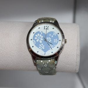May include: A silver-toned wristwatch with a light blue heart-shaped face adorned with white floral designs. The watch has a patterned green strap. The watch face displays the number 12 and black hands.
