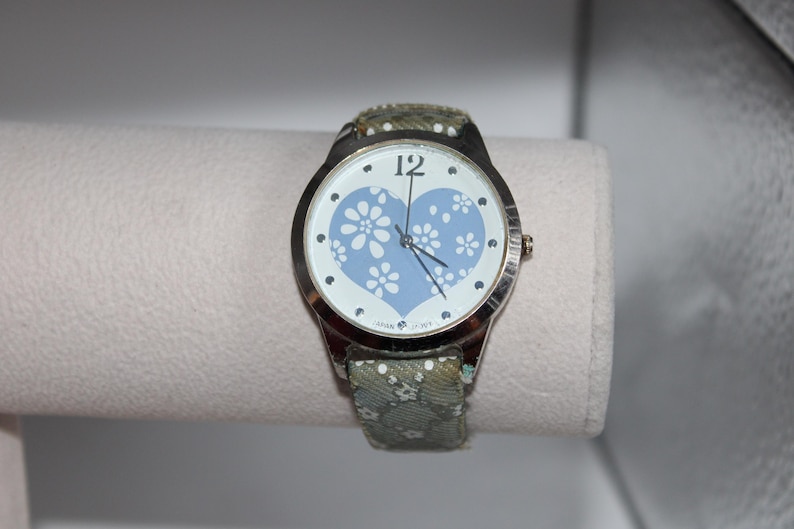 May include: A silver-toned wristwatch with a light blue heart-shaped face adorned with white floral designs. The watch has a patterned green strap. The watch face displays the number 12 and black hands.