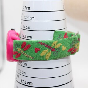 May include: A pink children's watch with a green fabric strap decorated with dragonflies and hearts. The watch is shown on a white cylinder with centimetre markings. The strap is approximately 1.9 cm wide.