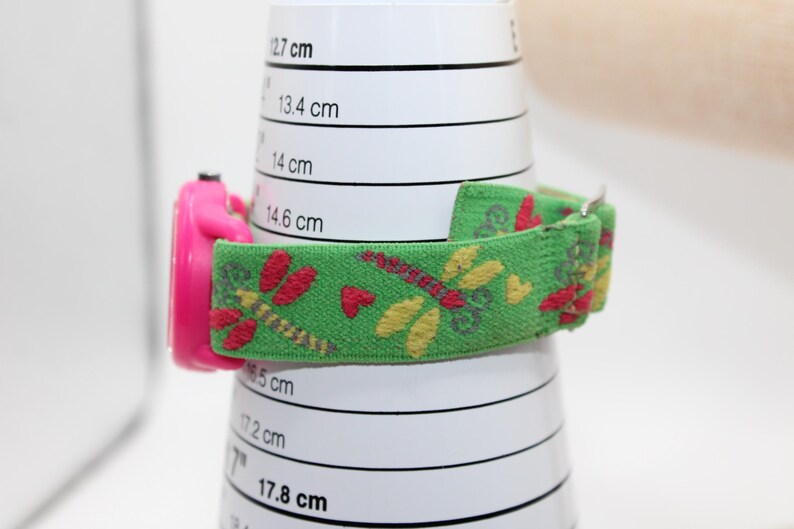 May include: A pink children's watch with a green fabric strap decorated with dragonflies and hearts. The watch is shown on a white cylinder with centimetre markings. The strap is approximately 1.9 cm wide.