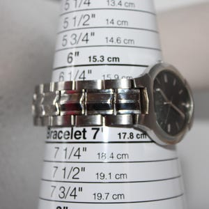 May include: A silver wristwatch with a black face is placed on a bracelet sizing tool. The tool shows measurements in inches and centimeters. The watch band is approximately 7 inches (17.8 cm) in circumference.
