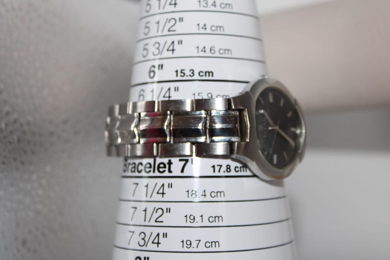 May include: A silver wristwatch with a black face is placed on a bracelet sizing tool. The tool shows measurements in inches and centimeters. The watch band is approximately 7 inches (17.8 cm) in circumference.