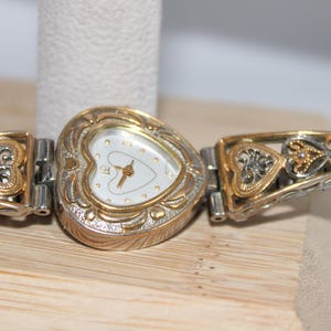 May include: A heart-shaped watch with a gold-tone bezel and a white face. The watch band features gold and silver-tone filigree and heart-shaped accents. The watch is displayed on a light-colored wooden surface.