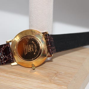 May include: A gold-plated watch with a black leather strap and a brown patterned accent. The watch face is visible, with the brand name "IMAGE" and other text. The watch is displayed on a wooden surface.