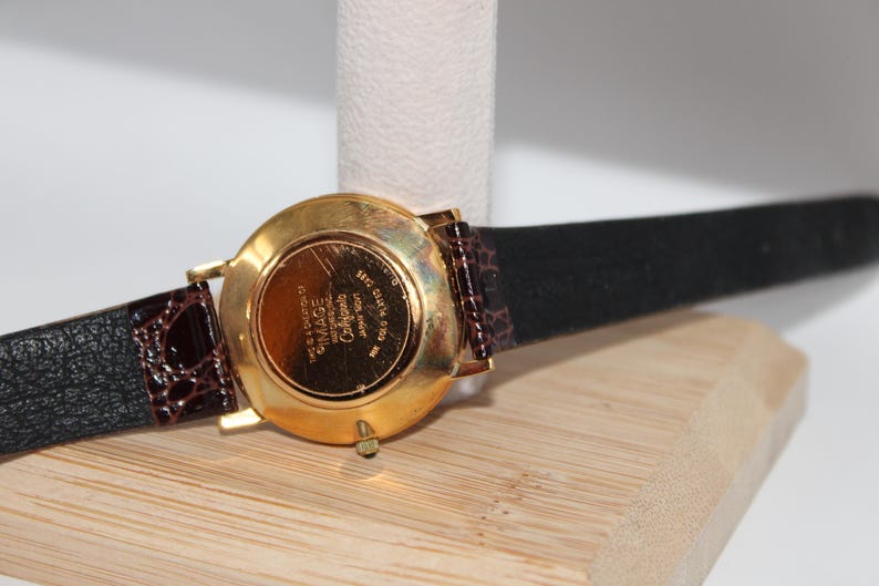 May include: A gold-plated watch with a black leather strap and a brown patterned accent. The watch face is visible, with the brand name "IMAGE" and other text. The watch is displayed on a wooden surface.