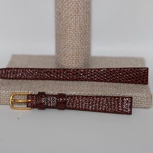 May include: A dark burgundy watch strap featuring a textured, lizard-like embossed pattern. The strap consists of two pieces, with the shorter section including a gold-toned metal buckle and two matching leather keepers for secure fastening.