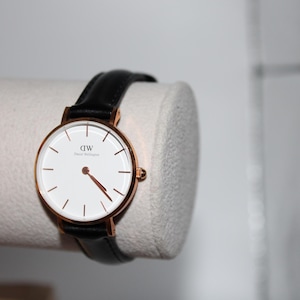 May include: A rose gold wristwatch with a white face and black leather band. The watch has a minimalist design with thin black hour markers and red hands. The watch is displayed on a white cylindrical object.
