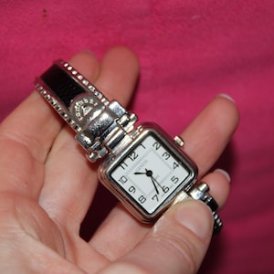 May include: Silver-toned wristwatch with a square white face and black hands. The watch has a black leather band with silver accents and a decorative clasp. The watch is held in a hand against a pink background.