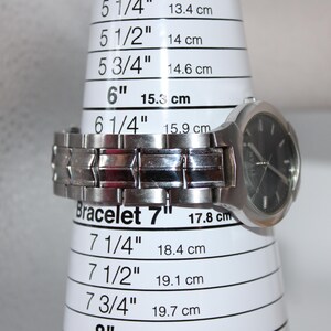 May include: A silver wristwatch with a black face is displayed on a white bracelet sizing cone. The cone shows measurements in inches and centimeters, with the watch resting at the 6-inch (15.3 cm) mark. The watch band is made of linked metal.