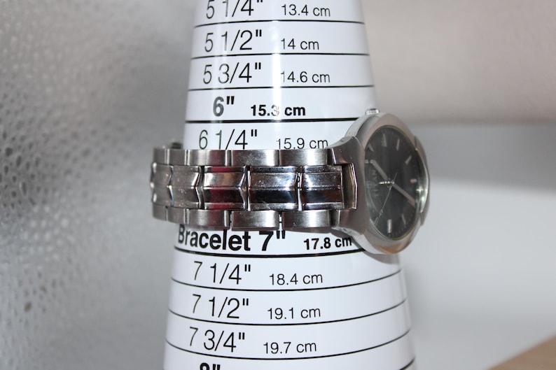 May include: A silver wristwatch with a black face is displayed on a white bracelet sizing cone. The cone shows measurements in inches and centimeters, with the watch resting at the 6-inch (15.3 cm) mark. The watch band is made of linked metal.