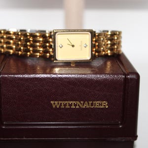 May include: A gold-tone Wittnauer wristwatch with a rectangular face, a cream-colored dial, and two small diamond markers at the three and nine o'clock positions. The watch features a linked gold-tone metal bracelet and rests on a dark brown box.