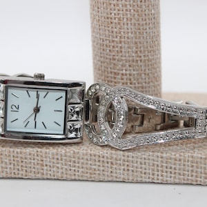 May include: Silver-toned rectangular watch with a white face and black hands. The watch features crystal accents around the face and on the band. The band is also silver-toned and has a decorative design.