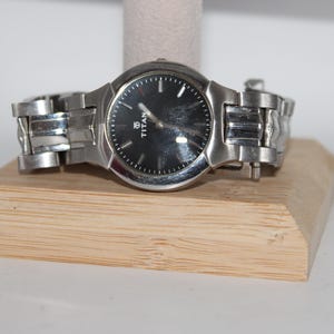 May include: A silver-toned wristwatch with a black face, resting on a wooden display stand. The watch features a round dial with silver hands and markers. The band is made of linked metal. The watch is a Titan brand.