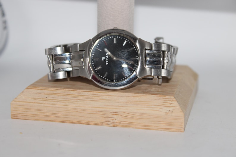 May include: A silver-toned wristwatch with a black face, resting on a wooden display stand. The watch features a round dial with silver hands and markers. The band is made of linked metal. The watch is a Titan brand.