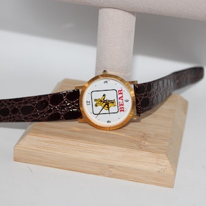 May include: A gold-toned wristwatch with a white face featuring a yellow bear graphic and the word "BEAR" in red. The watch has a brown, textured leather band and is displayed on a wooden stand.