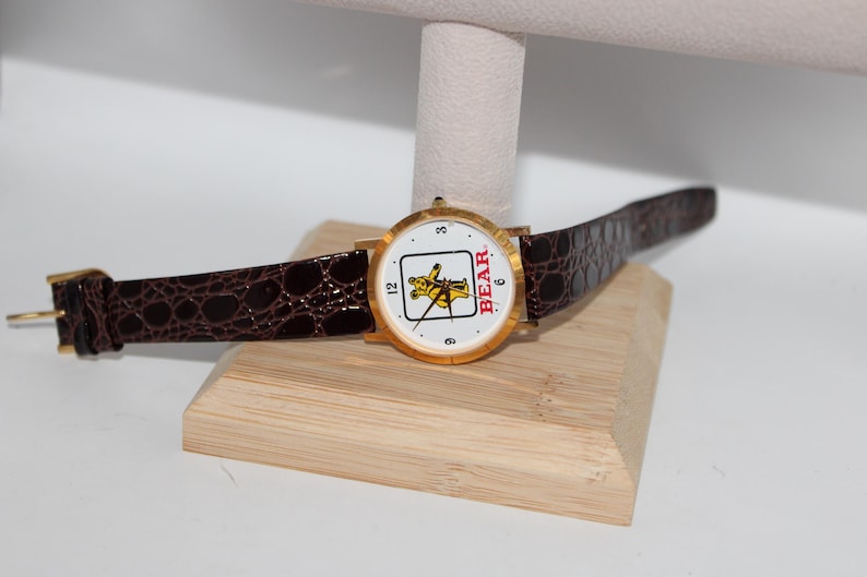 May include: A gold-toned wristwatch with a white face featuring a yellow bear graphic and the word "BEAR" in red. The watch has a brown, textured leather band and is displayed on a wooden stand.