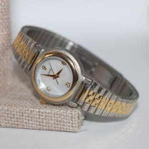 May include: A silver and gold-tone wristwatch with a white face and gold hands. The watch has a gold bezel and a silver and gold-tone stretch band. The watch is displayed on a beige textured surface.